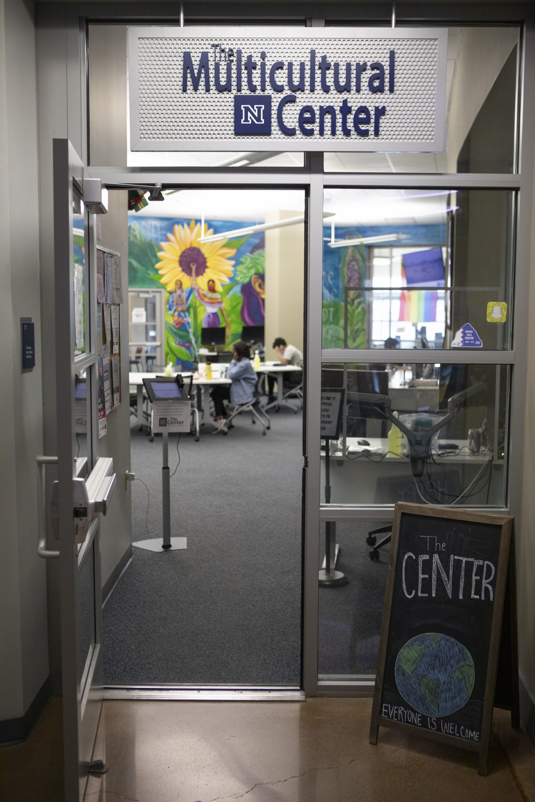 Open glass door with a silver sign above that read "The Multicultural Center" in blue letters with a UNR logo beneath it. Blurred sunflower mural in background of the image through the open door.