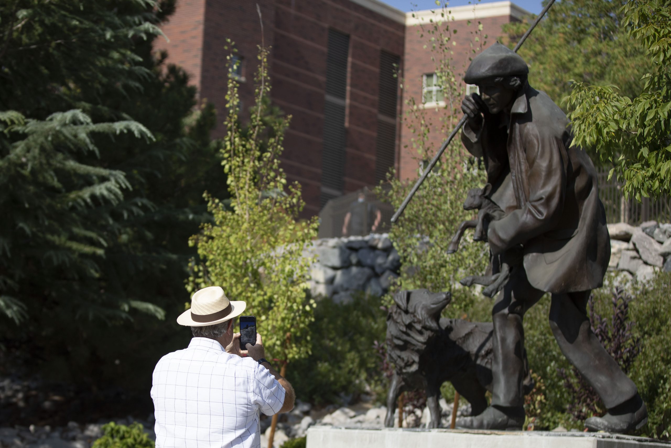 Statue of man with a sheep in his arms and a dog on his left on the right side of the image. A man in a tan hat wearing a blue shirt takes a photo of the staues on the left side.