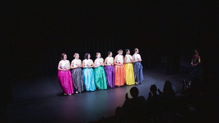 The girls in different colored long skirts stand in a line as they bow for the audience's applause on stage.