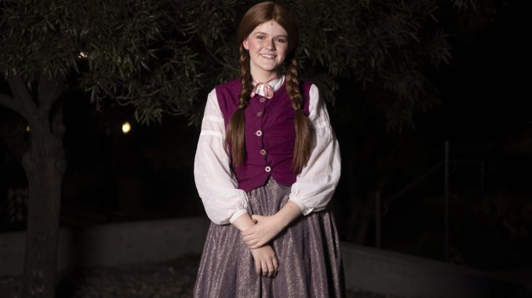 Madi Allen, a young girl stands outside against a dark background in her old fashioned costume of a purple tunic and white fluffy blouse underneath, double red braids, her arms crossed in front over her lighter purple tinted skirt.