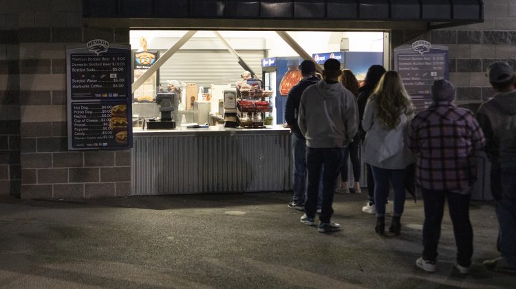 People standing in a line in front of a snack bar with the menu on the outside and a light which covers the inside of the mini shack.