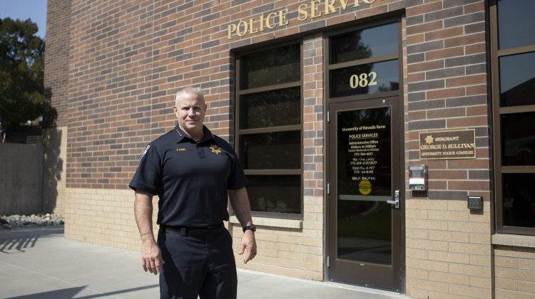 Man wearing all black with a grey buzzcut stands in front of a brick building with the words "police services" written on top in gold letters.