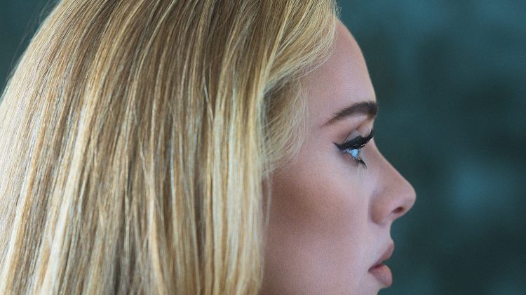 The artist Adele, posts with her right side profile face the camera against a blue background. Her hair is blonde and she is posing straight-faced with makeup on.
