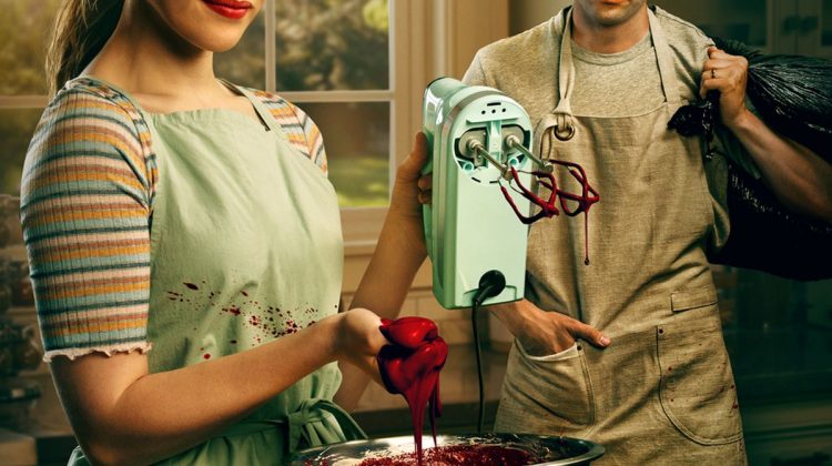 The main characters looking evil in a nice family home with aprons on. The woman is using a cake mixer to mix a bowl of blood while the man holds a knife and both stare at the camera.