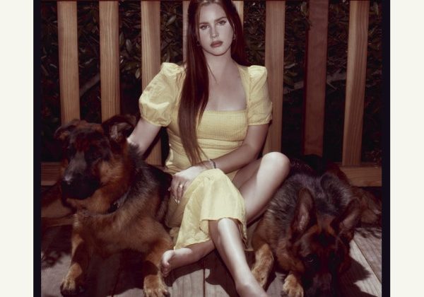 Lana Del Rey sits in between two dogs in a yellow dress, against a wooden railing with her album title and name at the top.