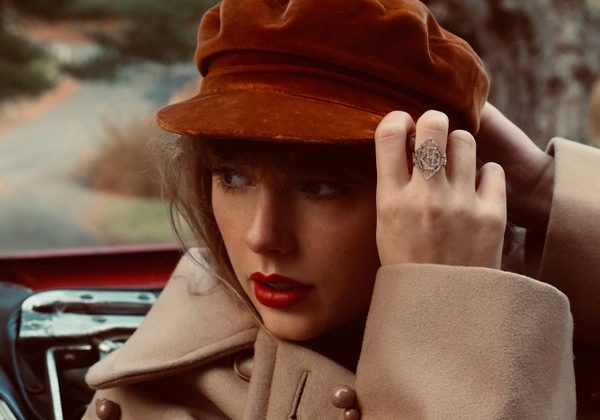 Taylor Swift poses with her hats on her red hat wearing a tan coat against a nature background.