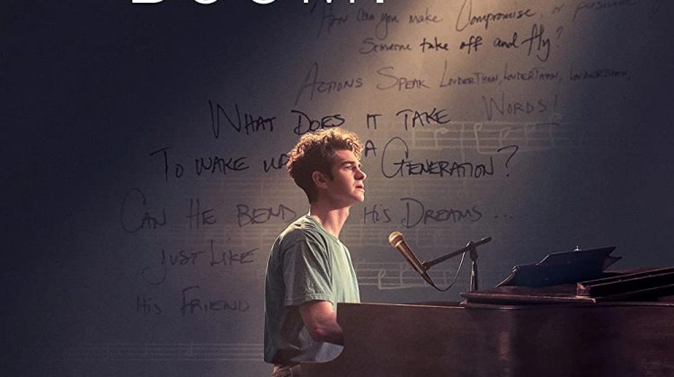 Andrew Garfield sits at a piano under a spotlight while script lyrics sit on a blank wall under a shadow behind him.