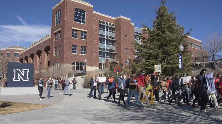 A group of people walk in front of a large birck building and the UNR logo