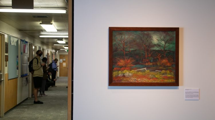A painting by Richard Guy Walton sits on a white wall while on the other side of the wall are a selection of students waiting for their class in a bright hallway.
