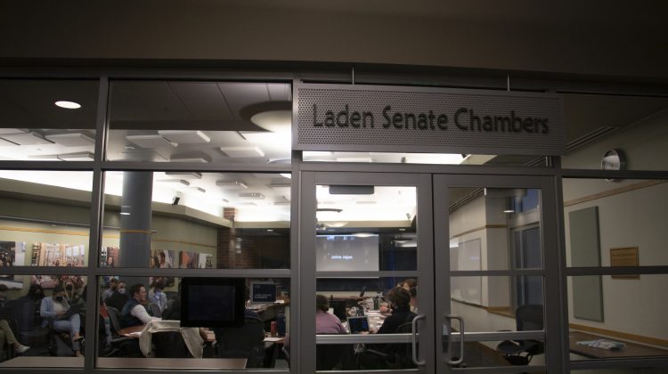 A sign that reads Laden Senate Chambers with a glass wall behind it.