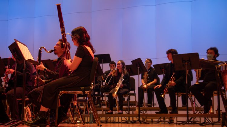 The concert winds are sitting, focused on their music against a blue and pink background.