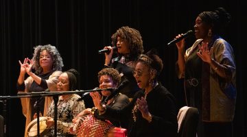 Six of the women in the group stand close together in a circle, singing their hearts out. The background is dark and they are all facing the audience.